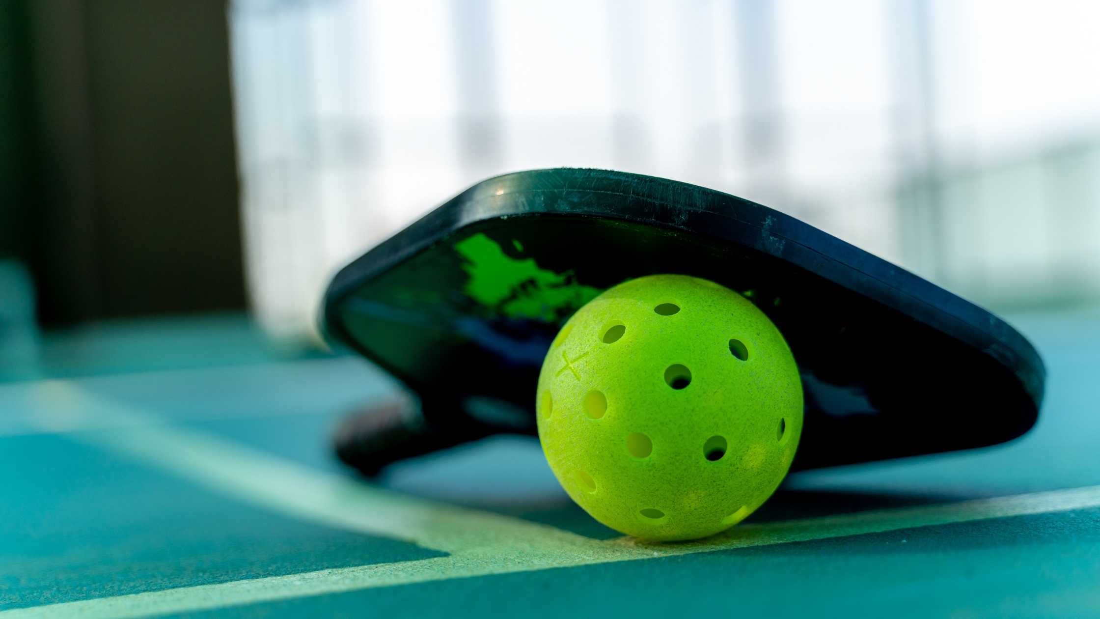 A close-up of a pickleball paddle resting on a court with a bright green pickleball underneath representing playing pickleball with the new official pickleball rules in 2025.