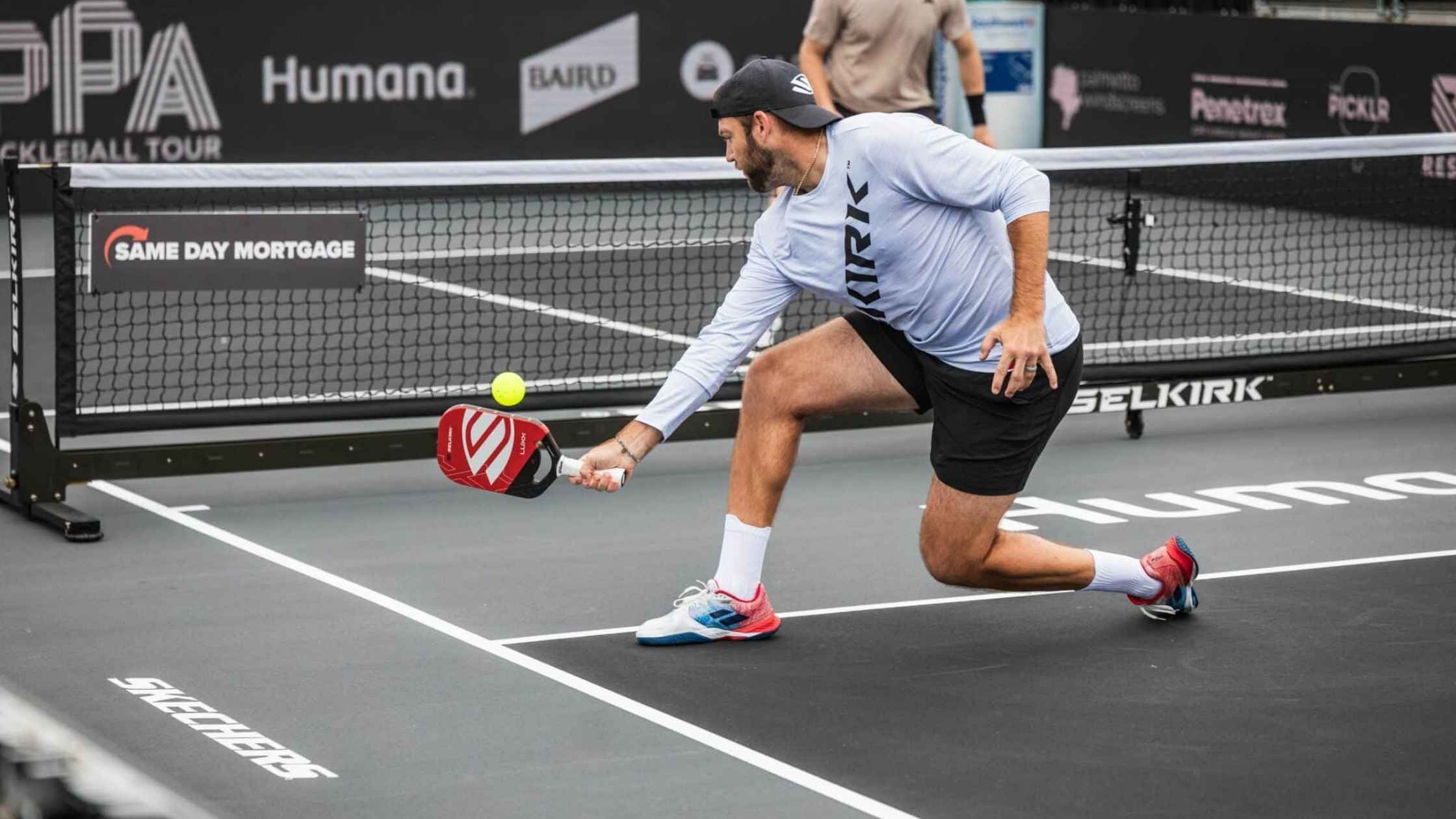 Pickleball player lunging to hit a low shot with a red Selkirk paddle during a singles match.